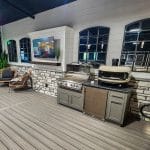 An outdoor kitchen setup featuring a gas grill, pizza oven, cabinetry, and a sink. Adjacent are two wicker chairs, a flat-screen TV mounted above, and plants along the wall.