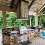 An outdoor kitchen features a stone and brick grill setup with stainless steel appliances, a wooden ceiling, and a view of surrounding trees and a pool.