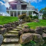 A two-story house with a stone facade and balcony is viewed from below. A custom porch in Glen Mills complements the architecture as a stone pathway and steps lead up a grassy hill to the house. The sky is partly cloudy.