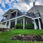 A custom porch in Glen Mills adorns this two-story house, boasting a large covered balcony framed by white pillars, all perched on a well-maintained grassy slope with rocks dotting the foreground. The sky overhead is partly cloudy.