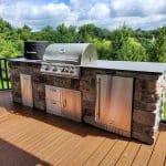 An outdoor kitchen on a custom hillside porch in Glen Mills featuring a stainless steel gas grill, stone countertops, and storage cabinets. The kitchen is set on a wooden deck overlooking a lush, green backyard.