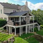 A large two-story home with light-colored siding features a spacious raised patio with stone detailing and a black railing, supported by white columns. The custom hillside porch in Glen Mills overlooks the backyard, which has a green lawn and a wooden fence.