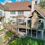 Two-story house with a stone chimney and a custom deck in Reading, featuring multi-level wood decks and outdoor seating areas. Manicured garden with stone steps and a green lawn surrounding the property. Bright, sunny day.