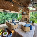 A spacious patio features wicker furniture with blue cushions, a stone fireplace with a TV above it, a wooden ceiling with fans, and views of lush greenery.