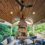 A covered outdoor patio features a stone fireplace, comfortable seating, ceiling fans, and overhead heaters. The area is surrounded by lush greenery.