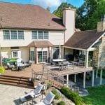 A two-story home with a large custom deck in Reading, featuring patio furniture, a hot tub, and a stone chimney. The backyard includes greenery and lounge chairs on a concrete patio.