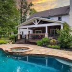 Backyard with a two-story house featuring a covered deck, surrounded by lush greenery and a large swimming pool with a built-in hot tub.