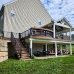 A two-story house with an outdoor deck and patio area. The deck includes a covered section with outdoor furniture and a ceiling fan. There is a staircase leading from the deck to the grassy backyard.