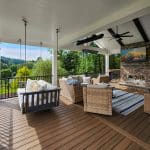 A covered patio with wicker furniture, a hanging bed swing, a fireplace, and a TV. The patio overlooks a grassy landscape with trees in the distance.