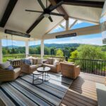 A covered outdoor patio features wicker furniture, a striped rug, a ceiling fan, and a TV mounted near a stone wall, overlooking a scenic view of grassy hills and trees.