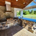 Outdoor kitchen with stainless steel grill, stone countertops, herringbone backsplash, and overhead wood paneling. Greenery and wrought iron fence visible through window.