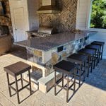 Outdoor kitchen with a stone island bar, six wicker bar stools, stainless steel grill, and a herringbone backsplash. The area features a fireplace with a flat-screen TV and wicker seating.