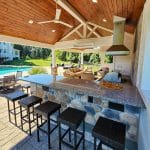 A covered outdoor kitchen with a stone bar countertop and four wicker bar stools faces a seating area and swimming pool. The space features wooden ceiling panels and a ceiling fan.