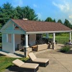 Outdoor gazebo with a covered lounge area featuring cushioned sofas, a built-in kitchen with a grill, and a patio with sun loungers. The background includes trees and a fenced yard.