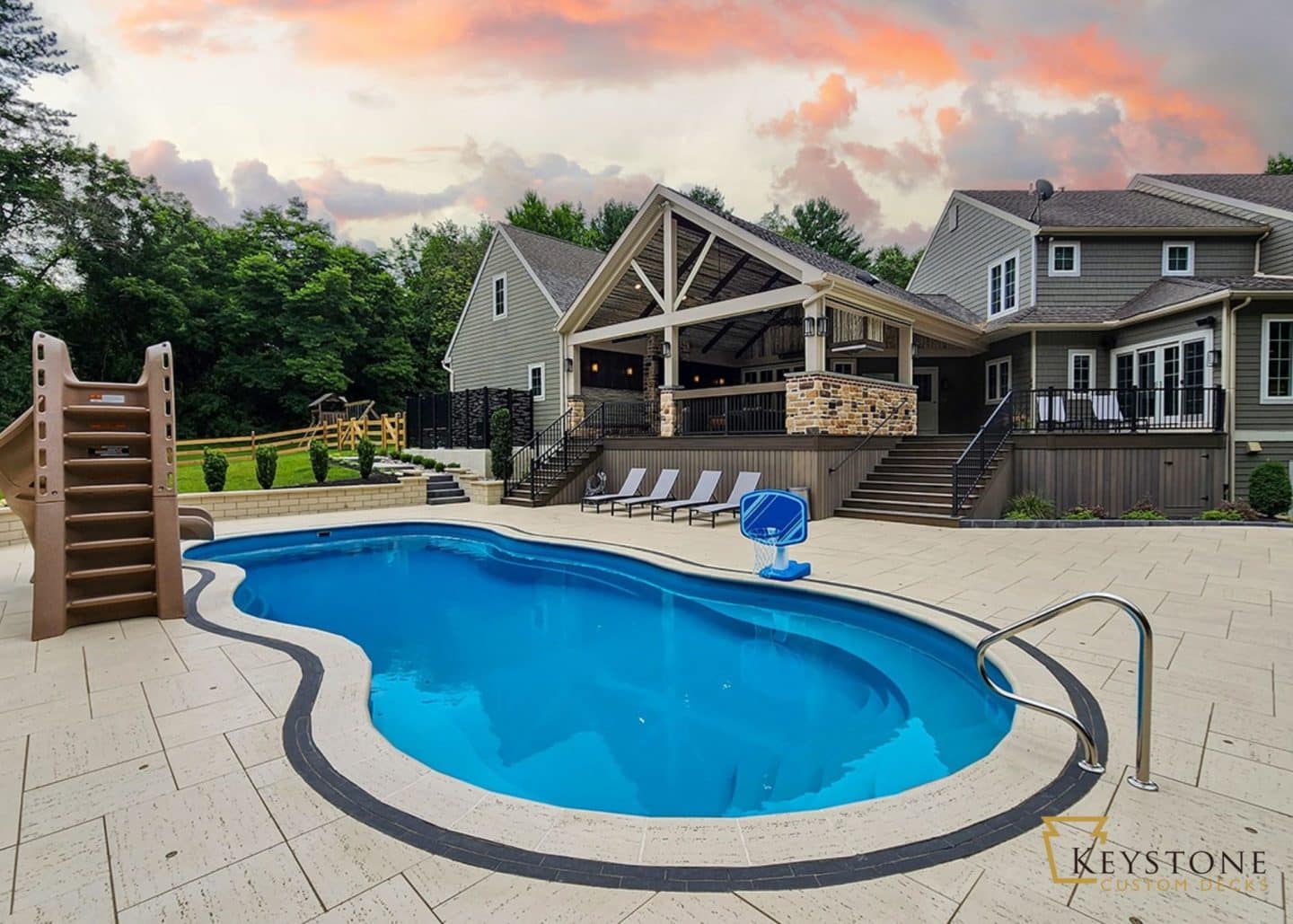 A backyard with a swimming pool, slide, and lounge chairs. The pool area is surrounded by a patio with a covered outdoor seating area, perfect for enjoying the outdoor living space. The house in the background has multiple levels and a large deck.