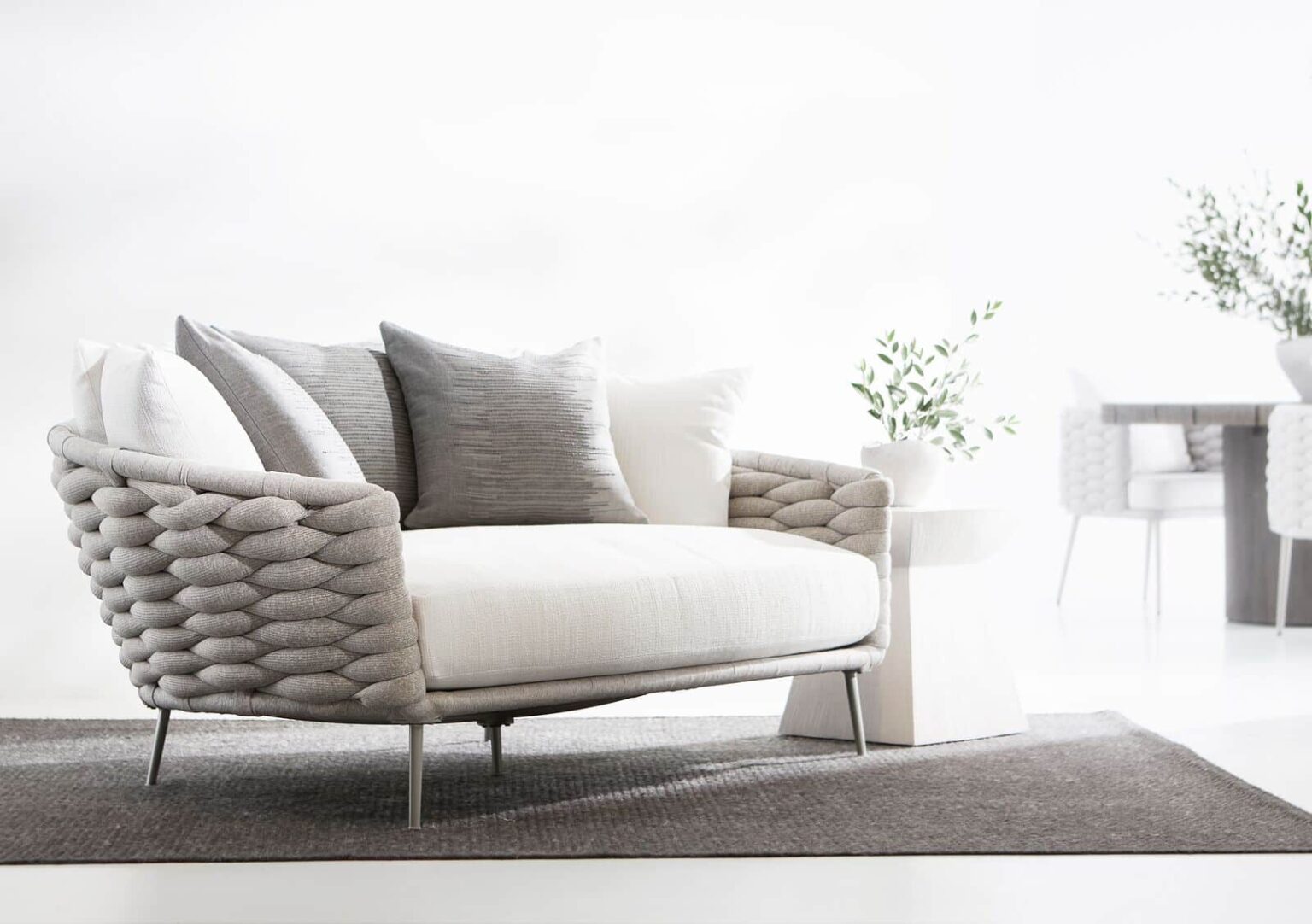 A modern, woven-texture loveseat with white and grey cushions, placed on a grey rug in a light, minimalistic room. A small white side table with a plant is positioned next to it.