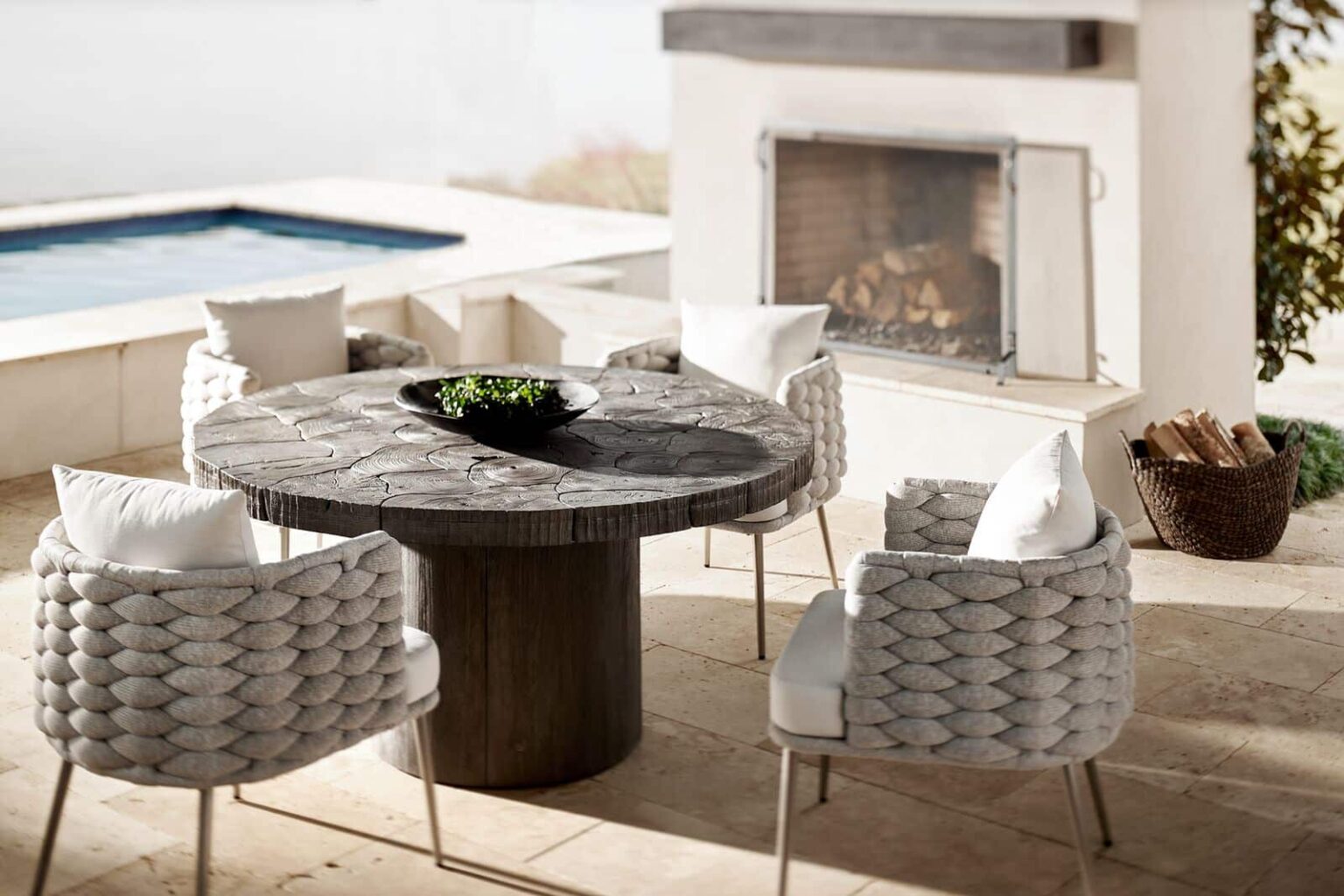 Outdoor patio with round wooden table, four cushioned chairs, a fireplace with stacked wood, and a pool in the background.