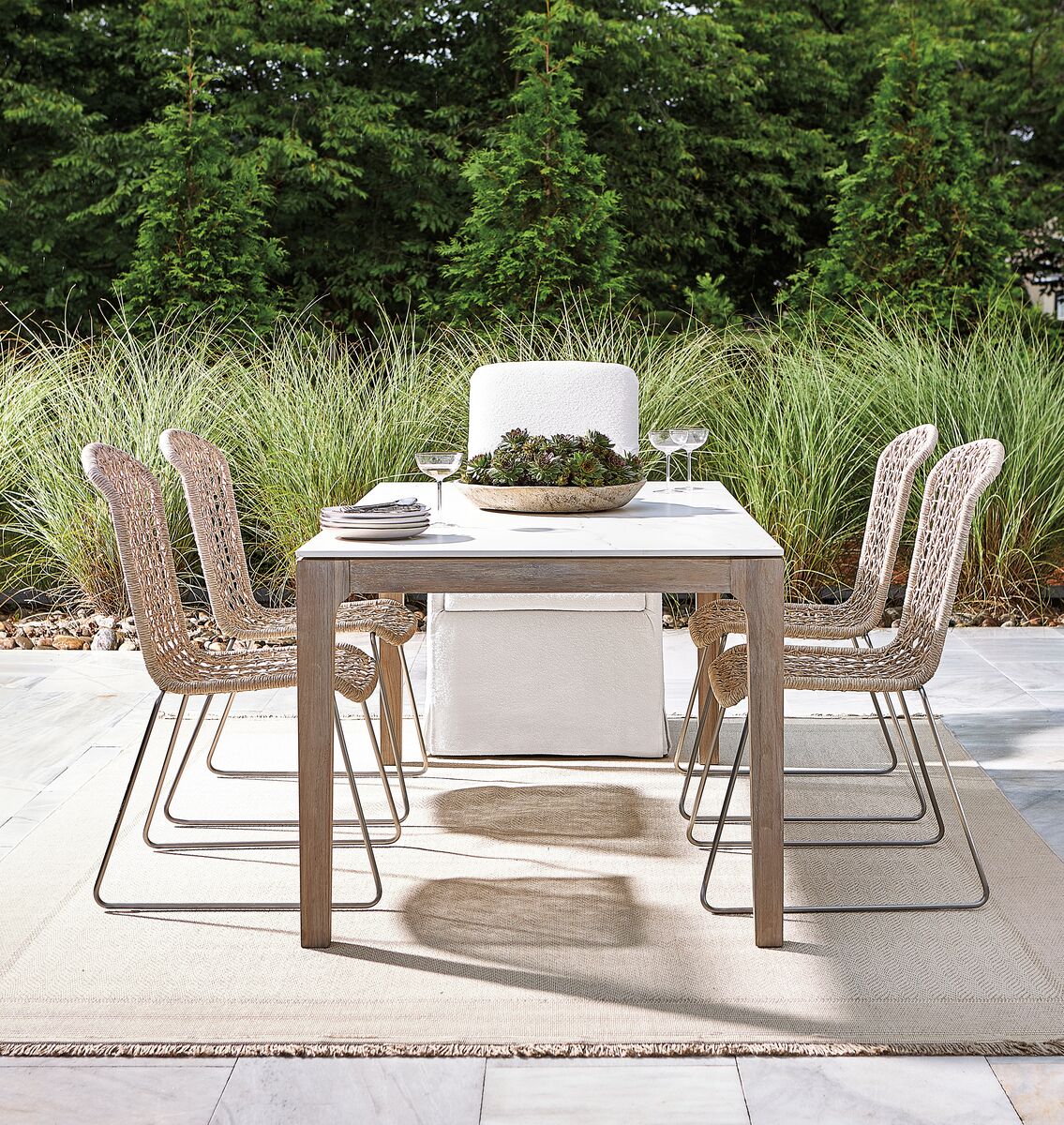 Outdoor dining set with six woven chairs and a rectangular table on a beige rug. A plant centerpiece and glassware are on the table. Green foliage is in the background.