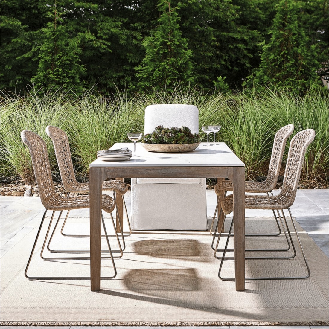 Outdoor dining setup with a rectangular wooden table, six wicker chairs, a central plant arrangement, and wine glasses against a backdrop of greenery.