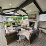 Covered outdoor patio with seating area including a sofa, chairs, and small tables. There is a fireplace with a mounted TV above it. The area overlooks a suburban neighborhood.