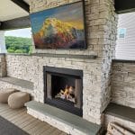 Outdoor seating area with a stone fireplace and a mounted TV displaying a mountain scene. Firewood is stacked to the right of the fireplace. Two round poufs and greenery are visible in the background.