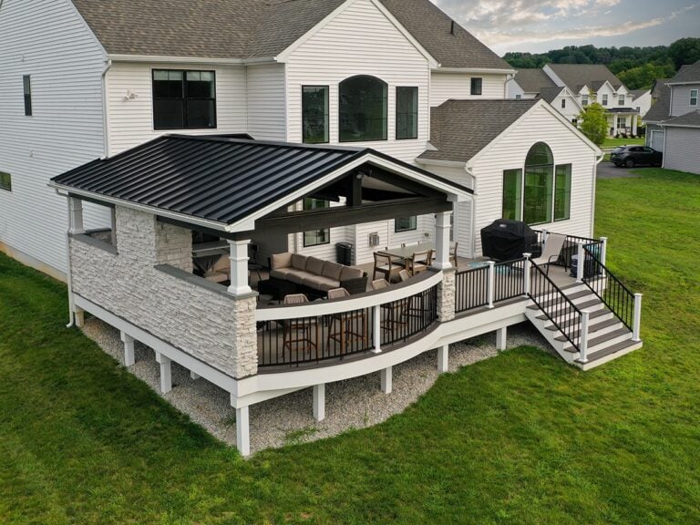 A two-story white house with a covered patio featuring outdoor seating, a grill, and stairs leading down to a grassy yard. The patio has a black metal roof and stone accents.
