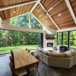 Spacious living room with wooden ceiling, large windows, and a view of a lush green yard. Features a sectional sofa, dining table, fireplace, and ceiling fan. Natural light fills the room.