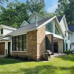 A modern two-story house with a mix of brick and white siding exterior, large windows, and an outdoor grill on the patio. The house has a sloped roof and is surrounded by trees and lush greenery.