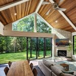 Modern living room with wooden ceiling, large windows, and glass folding doors opening to a green outdoor space. Features include a cozy sitting area, fireplace with mounted TV, and a long dining table.