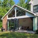 A modern house with a large open-concept room featuring floor-to-ceiling windows and a foldable glass door, leading to an outdoor area with a barbecue grill. The room contains a sofa and dining table.