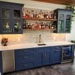 A home bar setup with blue cabinets, a sink, and shelving stocked with various liquor bottles. The counter holds a globe and a small fridge is built into the cabinetry on the left.