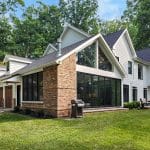 A modern two-story house with a mix of brick and white paneling exterior, large glass windows, and an attached garage set in a lush green yard with trees in the background.