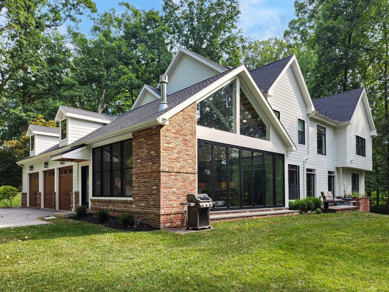 A modern two-story house with a mix of brick and white paneling exterior, large glass windows, and an attached garage set in a lush green yard with trees in the background.