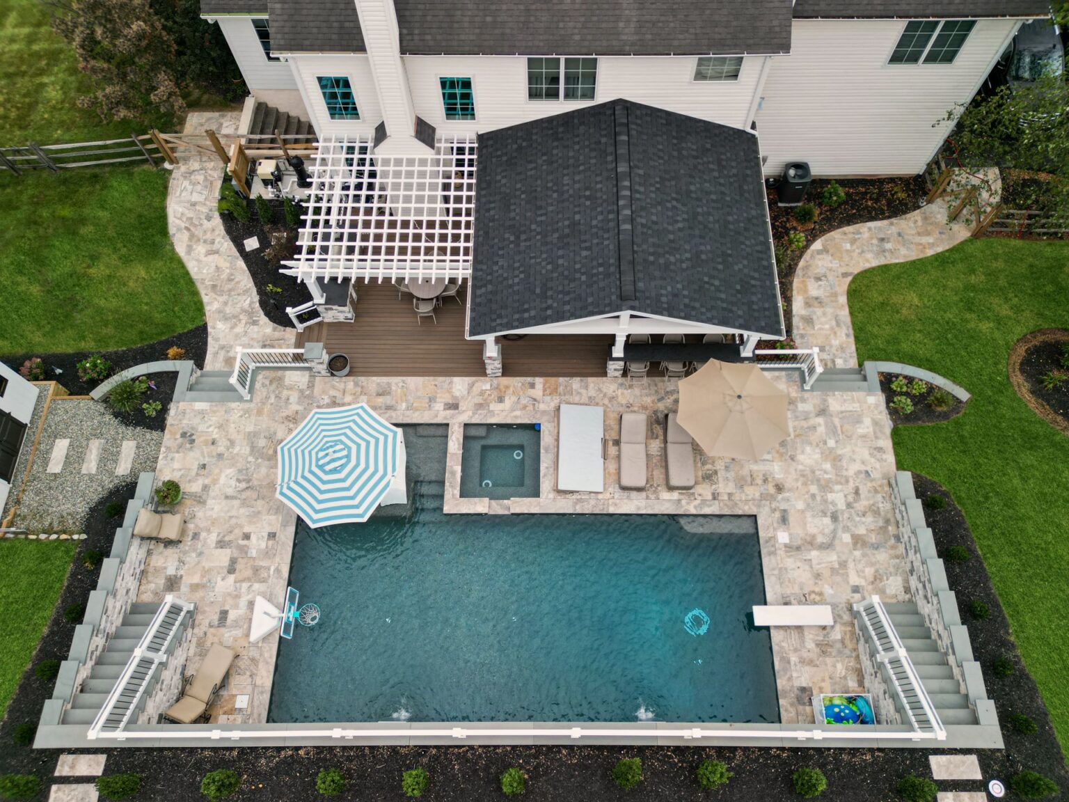 Aerial view of a backyard featuring a rectangular pool with a spa, surrounded by a stone patio with lounge furniture and umbrellas, near a two-story white house with a deck and pergola.