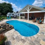 A backyard pool with a covered patio, seating area, and a stone deck. An RV and lawn are visible in the background. Bright, sunny day with a few clouds in the sky.