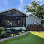 A modern house with a screened outdoor living area, adjacent deck, and well-maintained lawn. The garden features various plants and flowers, with a backdrop of trees and a clear sky.