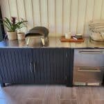 A modern outdoor kitchen with a black cabinet, stainless steel countertop, pizza oven, potted plants, wicker basket with towels, and a wooden tray with mugs.