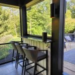 A cozy outdoor seating area with two bar stools at a high counter, surrounded by glass windows overlooking a lush green garden. A black speaker is placed on the counter.