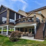 A multilevel house with an outdoor patio, stone columns, an upper deck with railing, screened area, wooden stairs, and lawn furniture on a grassy yard.