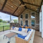 A covered patio with a high wooden ceiling features a seating area with a wicker couch and a dining table set. There's a barbecue grill near the stone wall with large windows.