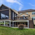 A large two-story house with a stone exterior features a covered patio, an upper deck with seating, and a wide staircase leading to a well-manicured lawn.