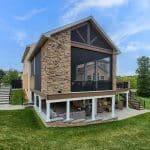 A two-story house with a stone exterior, elevated screened porch, and lower level outdoor seating area on a well-maintained lawn against a blue sky background.