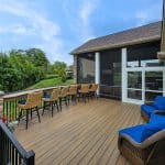 Spacious outdoor wooden deck with a row of bar-height chairs, wicker armchairs with blue cushions, and a view of a green lawn and trees.