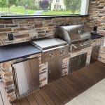 Outdoor kitchen with stainless steel appliances including a grill, side burner, and mini-fridge. The area features stone backsplash and countertops, and overlooks a grassy yard.