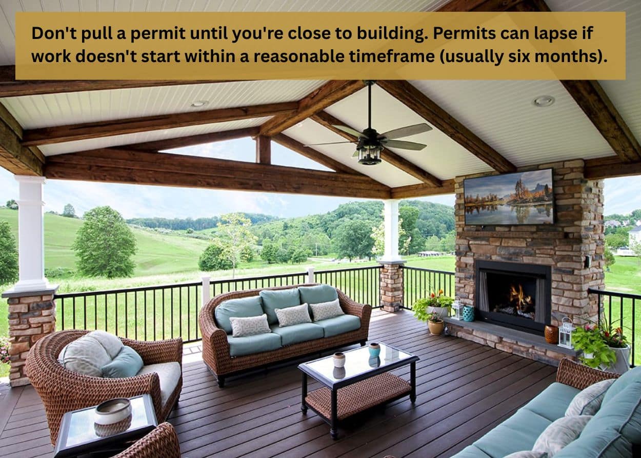 Covered deck with wood beams, ceiling fan, and stone fireplace overlooking open landscape.