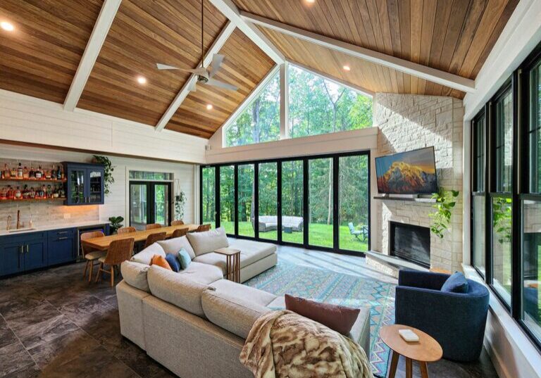 A modern living room with vaulted wooden ceilings, large windows, a sectional sofa, TV above the fireplace, built-in bar area, and dining table, overlooking a lush green backyard.