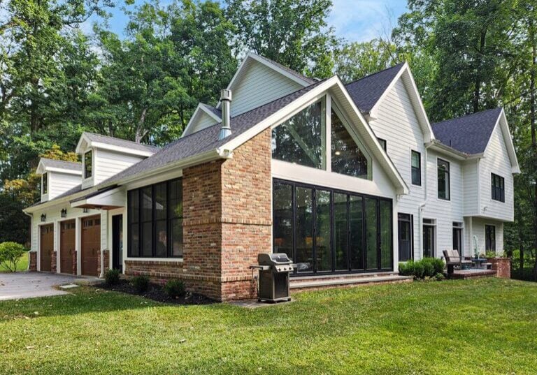 A modern two-story house with a mix of brick and white paneling exterior, large glass windows, and an attached garage set in a lush green yard with trees in the background.