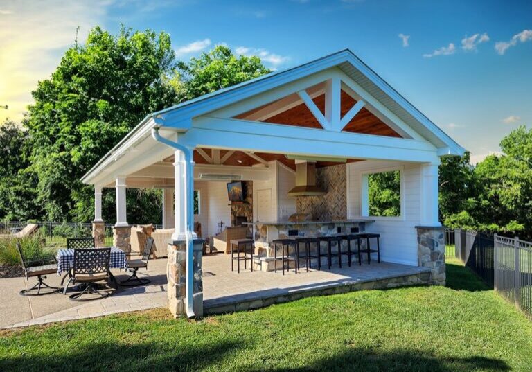 Outdoor pavilion with a gabled roof, featuring a kitchen area, bar seating, and patio furniture, set in a landscaped yard with trees in the background.