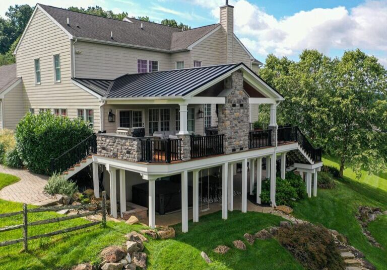 A large two-story home with light-colored siding features a spacious raised patio with stone detailing and a black railing, supported by white columns. The custom hillside porch in Glen Mills overlooks the backyard, which has a green lawn and a wooden fence.
