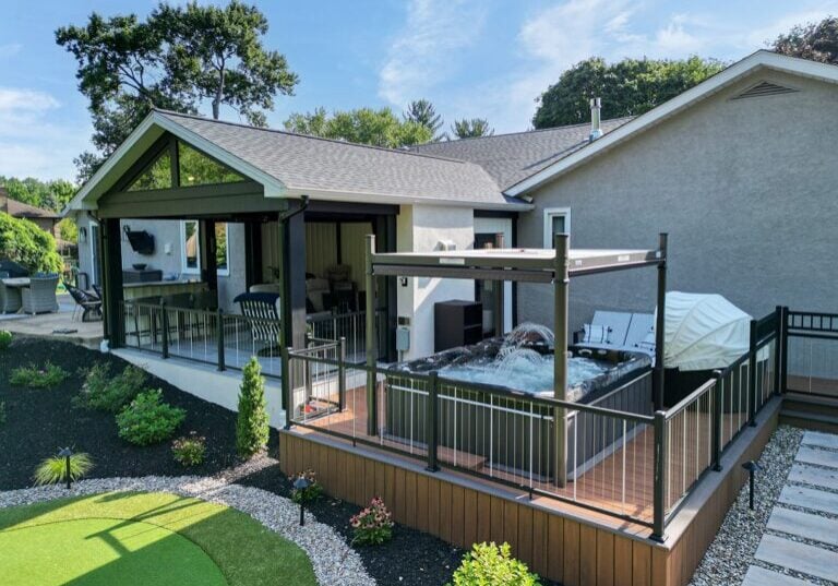 A modern backyard featuring a covered patio with seating, adjacent to a fenced-in hot tub area. The yard is landscaped with greenery and a small putting green. Houses and trees are visible in the background.